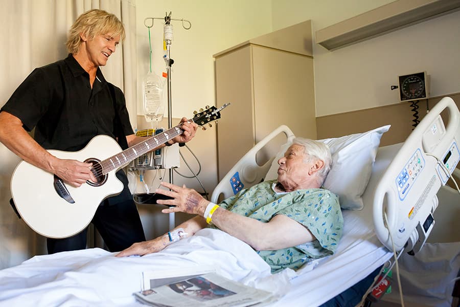 Musician playing acoustic guitar for patient in hospital bed during bedside performance