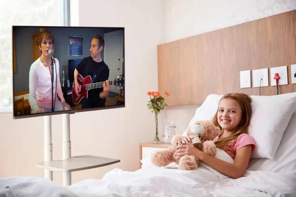 Patient in hospital bed watching live musicians perform via Zoom on bedside monitor