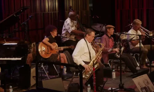 Professional jazz musicians performing live on stage with piano, guitar, saxophone, drums, and trumpet against red curtain